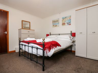A bedroom with a bed covered in white linens and a red throw a chest of drawers with a mirror a wardrobe and two paintings on the wall at Coach House Cottage in Bude