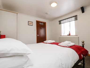 A bedroom with a bed made with white sheets and red blanket near a wooden door and window with plaid valance at Coach House Cottage in Bude