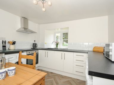 A kitchen with white cabinets black countertops a wooden table with two cups a stove a microwave a kettle and a window with flowers at Lee Barton Farmhouse in Bude