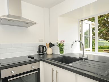 A kitchen with an oven stove kettle utensils flowers sink and an open window with a garden view at Lee Barton Farmhouse in Bude
