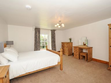 A bedroom with a wooden bed white bedding wooden dressers a window with curtains and a vanity at Lee Barton Farmhouse in Bude