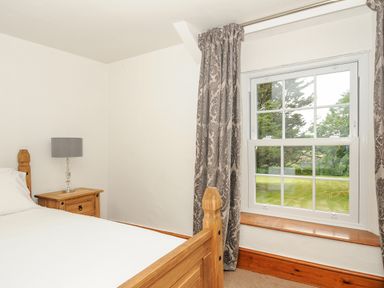 A bedroom with a wooden bed a bedside table with a lamp and a window with patterned curtains at Lee Barton Farmhouse in Bude