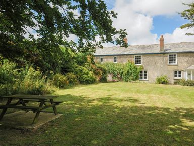 A garden with a wooden picnic table and grass in front of a stone farmhouse with climbing plants at Lee Barton Farmhouse in Bude