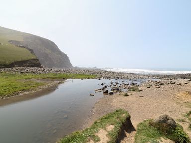 A rocky coastline with a small stream of water flowing towards the sea and hills in the background at Lee Barton Farmhouse in Bude