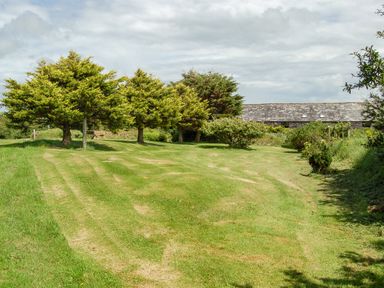 A grassy garden area with trees shrubs a bench and a long building in the background at The Roundhouse in Bude