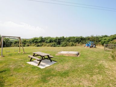 An outdoor playground area with a wooden picnic table two swings a sandbox and a blue toy tractor on grass at The Roundhouse in Bude