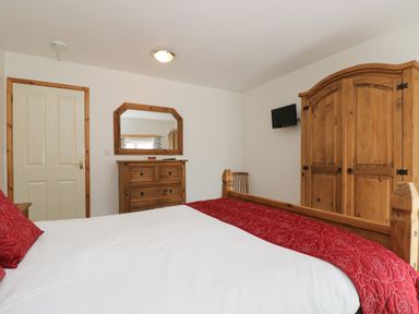 A bedroom with a wooden bed with red bedding a wooden wardrobe wooden dresser mirror and a wall-mounted tv at The Roundhouse in Bude