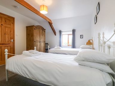 A bedroom with two single beds white bedding a wooden wardrobe a chair and a window with curtains at Swallow Cottage in Bude
