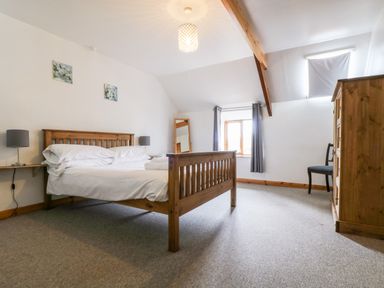 A bedroom with a wooden bed frame, two bedside lamps, a standing mirror, a window with curtains, a wooden wardrobe, and a chair at Swallow Cottage in Bude