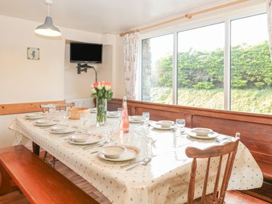 A dining room with a table set for eight people including plates bowls glasses and cutlery with a vase of flowers and a bottle with a large window showing greenery at The Wagon Linney in Bude