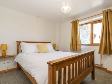 A bedroom with a wooden bed frame white bedding yellow cushions two yellow lamps and yellow curtains at The Wagon Linney in Bude