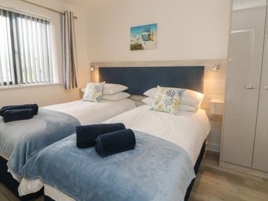 A bedroom with two single beds with blue throws pillows and towels a window with blinds and a wardrobe at Devonair in Westward Ho