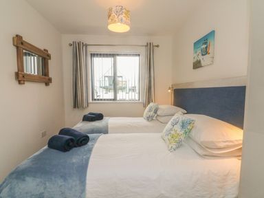A bedroom with two single beds blue throws and rolled towels a wooden mirror a window with striped curtains and a beach themed painting at Devonair in Westward Ho