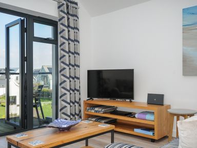 A living room with a wooden coffee table and TV unit next to a glass door with patterned curtains at Devonair in Westward Ho