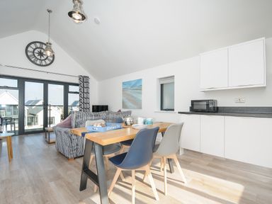 A dining area with table and chairs next to a kitchen and a living room with sofa and wall clock at Devonair in Westward Ho