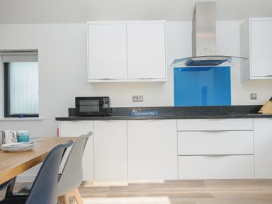 A kitchen with white cabinets black countertop a microwave a blue glass splashback and part of a wooden dining table with chairs at Devonair in Westward Ho