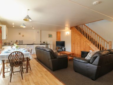 A living and dining area with a wooden dining table set with dishes and cutlery leather sofas a coffee table a television under stairs and kitchen appliances at The Dairy in Bude