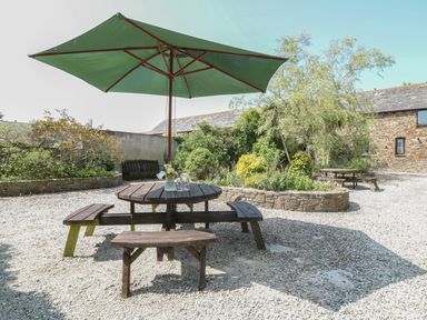 An outdoor area with wooden picnic tables and green umbrella surrounded by trees and stone walls at The Dairy in Bude