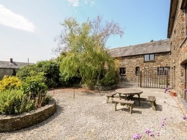 A courtyard with a round wooden picnic table and benches surrounded by stone buildings and plants at The Shippon in Bude