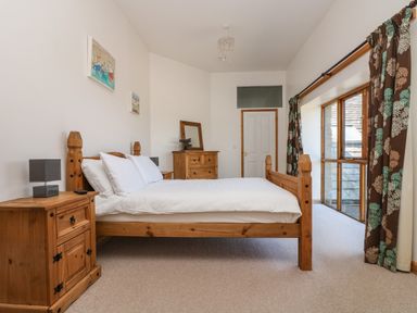 A bedroom with a wooden bed and matching bedside table and dresser with a window with curtains at The Shippon in Bude