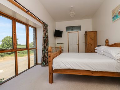 A bedroom with a wooden bed with white bedding a wooden wardrobe a chair and a wall-mounted TV with large windows and floral curtains at The Shippon in Bude
