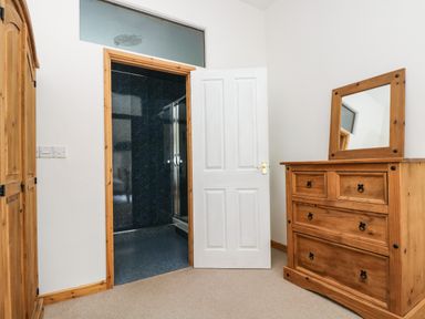 A bedroom area with wooden wardrobe and chest of drawers with mirror and a white door leading to a bathroom with dark walls at The Shippon in Bude