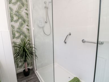 A shower area with white tiles chrome grab bars a handheld showerhead and a potted plant with leaf patterned wallpaper at Martingale in Torquay