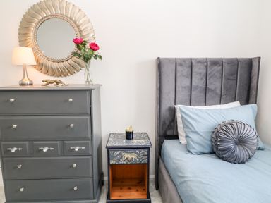 A bedroom with a gray dresser with a lamp and flowers a small decorated bedside table and a bed with a gray headboard and blue bedding at Martingale in Torquay