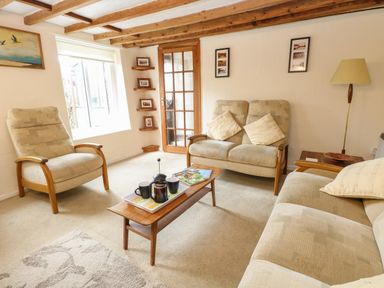A living room with beige armchairs a wooden coffee table with a tea set and magazines a standing lamp and a window at Primrose Cottage in Stiffkey