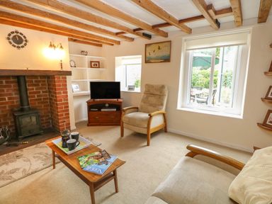 A living room with wooden beams on the ceiling a brick fireplace a television on a wooden stand an armchair a sofa a wooden coffee table with cups and magazines and windows showing a garden at Primrose Cottage in Stiffkey