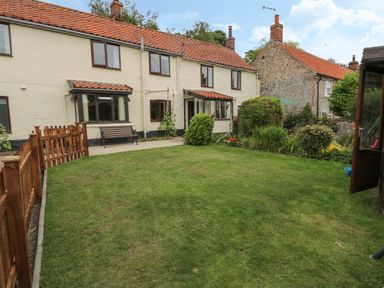 A garden with lawn, flowerbeds, and shrubs behind a row of terraced houses at Primrose Cottage in Stiffkey