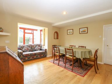 Dining room with a table covered by a green polka dot tablecloth surrounded by six wooden chairs and a floral patterned sofa by a window at Primrose Cottage in Stiffkey