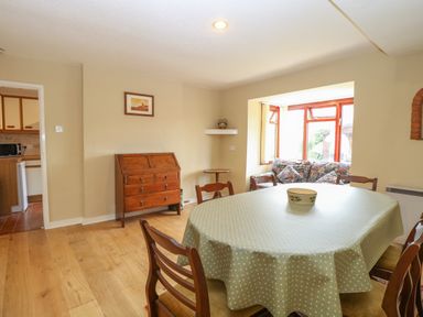 A dining room with a round table covered with a green polka dot tablecloth surrounded by wooden chairs a wooden chest of drawers and a window seat with cushions at Primrose Cottage in Stiffkey