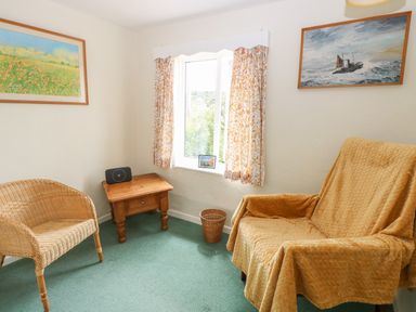 A small sitting room with wicker chair wooden side table and armchair with cover at Primrose Cottage in Stiffkey