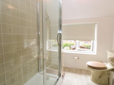 A bathroom with a shower enclosure beige tiles a toilet with a wooden seat and a window with a roller blind at Primrose Cottage in Stiffkey