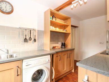 A kitchen with wooden cabinets washing machine utensils hanging on the wall and a clock at Primrose Cottage in Stiffkey