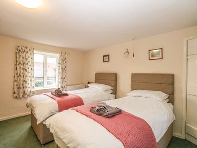 A bedroom with two single beds each with white bedding and pink throws near a window with floral curtains at Primrose Cottage in Stiffkey