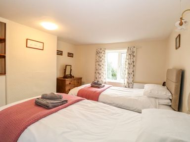 A bedroom with two single beds featuring pink throws and gray towels a wooden dresser a window with patterned curtains at Primrose Cottage in Stiffkey