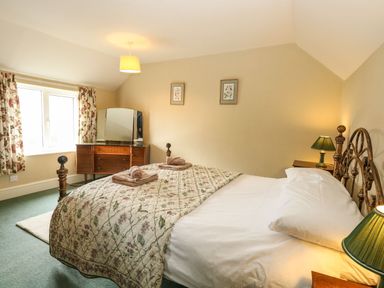 A bedroom with a double bed, nightstands with lamps, floral curtains, a dresser with a mirror and framed pictures on the wall at Primrose Cottage in Stiffkey