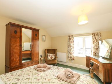 A bedroom with a bed, wooden wardrobe, armchair, dressing table, and window with floral curtains at Primrose Cottage in Stiffkey