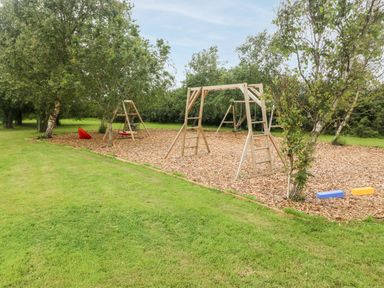 An outdoor playground area with wooden swing sets and a slide surrounded by trees on a grassy field at Llofft Yr Yd in Llandwrog near Dinas Dinlle