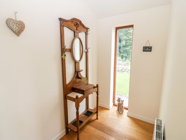 A hallway with a wooden hall tree featuring an oval mirror a small shelf and hooks next to a tall narrow window with a garden view at Llofft Yr Yd in Llandwrog near Dinas Dinlle