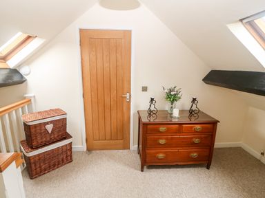 A hallway with a wooden door a wooden chest of drawers with candles and flowers and two wicker baskets stacked near a railing at Llofft Yr Yd in Llandwrog near Dinas Dinlle