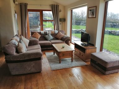 A sitting room with two sofas a coffee table a TV on a stand and large windows showing a garden at Llofft Yr Yd in Llandwrog near Dinas Dinlle