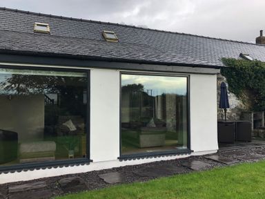 The exterior of a building with large windows showing indoor seating and a garden with patio furniture and a closed umbrella at Llofft Yr Yd in Llandwrog near Dinas Dinlle