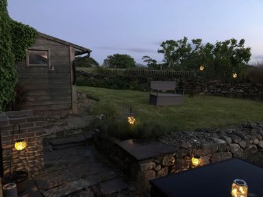 An outdoor garden area with stone paths a wooden shed a chair a tree with hanging lights and a stone wall at Llofft Yr Yd in Llandwrog near Dinas Dinlle
