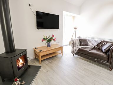 A living room with a wood burning stove a wooden coffee table with flowers and a brown leather sofa at 1 Mountain View in Talwrn near Llangefni