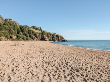 A sandy beach with footprints next to the sea and a tree covered cliff in the background at Yew Tree Cottage in Stoke Fleming