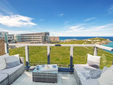 A balcony with wicker furniture and cushions overlooking a grassy area buildings and the sea at Breakers in Newquay