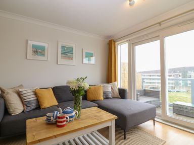 A living room with a gray sectional sofa yellow and striped cushions a wooden coffee table with a tea set and flowers near large windows at Breakers in Newquay